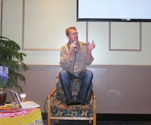Sex and relationship expert Reid Mihalko wearing a beige buttondown shirt and jeans sitting on the back of a chair speaking into a mic lecturing