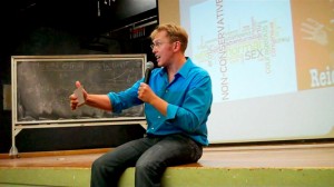Sex and relationship expert Reid Mihalko in a blue, button up shirt, sitting on an auditorium stage with hand held mic and note cards, lecturing with a slide show on the projector screen behind him