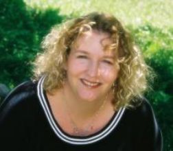 White woman with curly blonde hair, sitting outside, smiling at the camera, wearing a dark blue blouse with white stripes across the scoop neck.