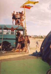 Thirteen women and Reid Mihalko gathered around a blue bus at Burning Man after erecting a rainbow pride flag and a yellow flag with a beaver and "Beaverton" painted on it.
