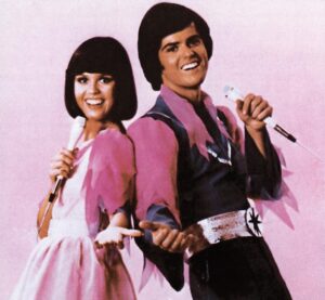 Young Donny and Marie Osmond standing back to back with mics in their hands smiling at the camera