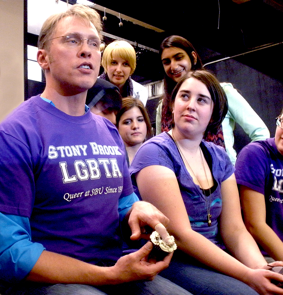 Sex and relationship educator Reid Mihalko of ReidAboutSex.com wearing a purple Stony Brook LGBTA t-shirt sitting with Stony Brook College students teaching a safer sex demonstration on a cupcake wearing a finger cot