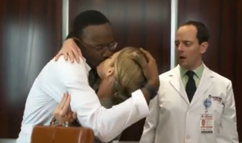 Inside an elevator, a man and a woman in lab coats kiss passionately while another man in a lab coat stares in shock.