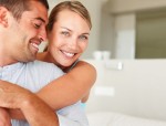 A smiling, blonde haired, blue-eyed woman embraces her handsome, smiling partner from behind as she looks to the camera.