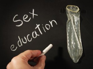 a teacher's hand writes the word "sex education" on a chalk board which has an unrolled condom taped to it.