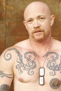 Head and shoulders photo of sex educator and transexual adult performer Buck Angel wearing military dog tags and no shirt, showing off his tatoos