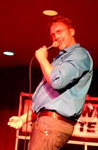 Sex and relationship expert Reid Mihalko wearing a blue shirt and jeans, pantomiming masturbating with one hand and speaking into a microphone with the other on stage during a Bawdy Storytelling event