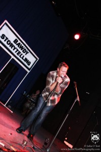 Sex and relationship expert Reid Mihalko wearing a brown, plaid shirt and jeans on stage huddling around the microphone with a look of pain on his face and the Bawdy Storytelling banner behind him