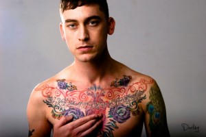Sex educator and queer blogger Brandon B posing without a shirt, hand over his heart, colorful chest tattoos exposed
