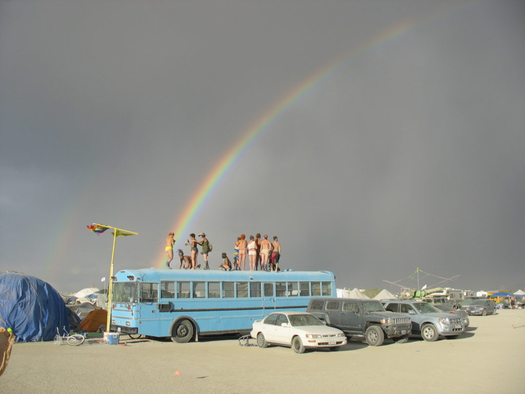 Image from Burning Man of a few dusty cars parked in front of a blue school bus with a group of people standing on the roof with a large rainbow in the sky above it