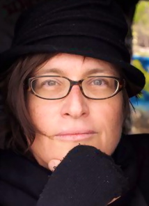 A close up of author and lecturer Susie Bright contemplatively facing the camera wearing a black hat, thick rimmed glasses and black fingerless gloves 