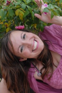 Healer and teacher Monique Darling with a huge, happy, laughing smile and flowing brown hair in a flowery bush
