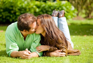 Couple in a green shirt and brown shirt kissing while laying on a green lawn