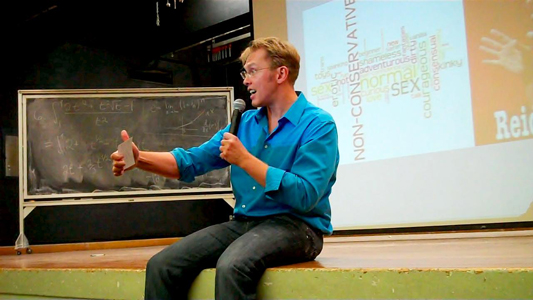 Sex and relationship expert Reid Mihalko in a blue, button up shirt, sitting on an auditorium stage with hand held mic and note cards, lecturing with a slide show on the projector screen behind him