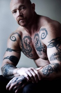 A tattooed Buck Angel seated and shirtless with his arms resting on his knees looking directly at camera