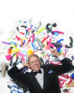 Sex and relationship expert Reid Mihalko of ReidAboutSex.com wearing a tux lying in a pile of sex toys