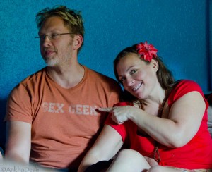 Sex educator Reid Mihalko of ReidAboutSex.com wearing an orange Sex Geek teeshirt and intimacy coach Monique Darling of DivineInterludes.com dressed in a beautiful red dress and red flower in her hair teaching together