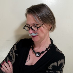 Sex educator Carol Queen posing for the camera in a black dress, arms akimbo, wearing a fake mustache