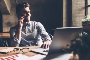 Confident young man in smart casual wear talking on the mobile phone and smiling while sitting at his working place in office