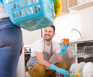 Young couple doing chores together. Man loading dishwasher and wife holding laundry basket