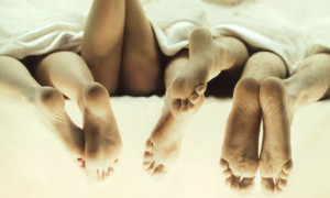Four pairs of adult feet in bed naked peeking out from under white sheets