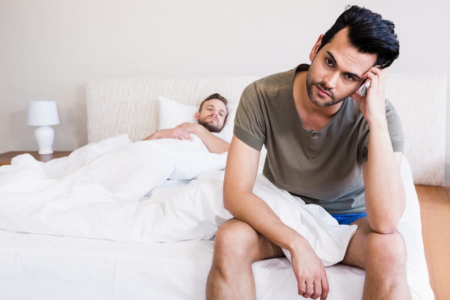 Thoughtful man sitting on bed while boyfriend sleeping