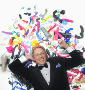 Sex and relationship educator Reid Mihalko of ReidAboutSex.com wearing a tuxedo, laying in a pile of sex toys against a white background