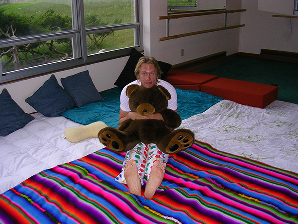 Cuddle Party creator and sex and relationship educator Reid Mihalko back in 2008 waiting for a Cuddle Party to begin, hugging a large teddybear sitting in pajamas on a rainbow blanket