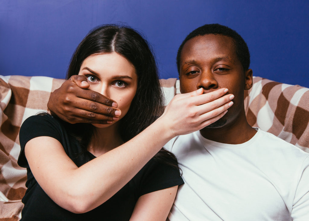 A close up of a couple sitting on a couch - a black man in a white t-shirt and caucasian woman in a black t-shirt - each holding their hands over the other's mouth in a "speak no evil" type pose