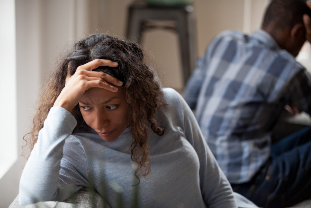 Tired, upset black woman feeling worried and her male partner in the background with his back turned to her.