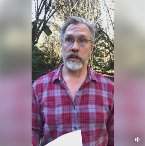 Image screen capture from Facebook Live of Reid Mihalko with grey and blonde beard, standing outside, wearing a red and grey plaid flannel shirt, holding some white paper, with trees in the background.