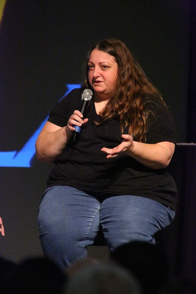 Cathy of TheIntimacyDojo.com sitting on a stage holding a microphone, speaking, wearing a black shirt and blue jeans, gesturing with her left hand.
