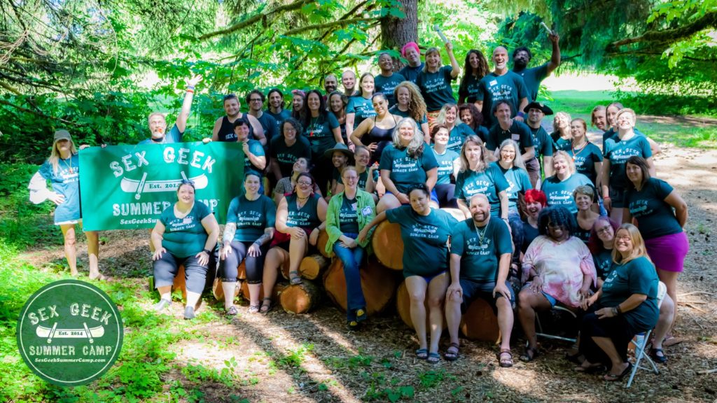 Group photo from Sex Geek Summer Camp 2019 of four dozen sex educators standing and sitting in the forest, some on large logs, many wearing green Sex Geek Summer Camp t-shirts, and a few holding up a Camp banner.