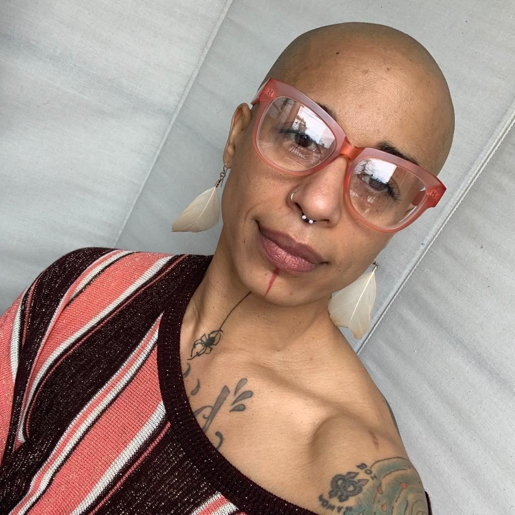 Image of Ignacio G. Hutía Xeiti Rivera, a Queer, Trans/Yamoká-hu/Two-Spirit, Black-Boricua, and Taíno activist, with a shaved head, wearing large framed, translucent, pinkish eyeglasses, white feather earrings, and a red and black striped blouse with one side of the neck draped over exposing their shoulder.