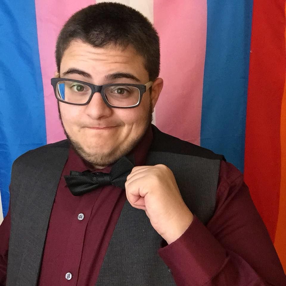 Photo of a white man with short brown hair with a playful smirk and raised eyebrows (Mason Luke) wearing glasses, and adjusting his bow tied with one hand standing  in front of a trans-pride flag