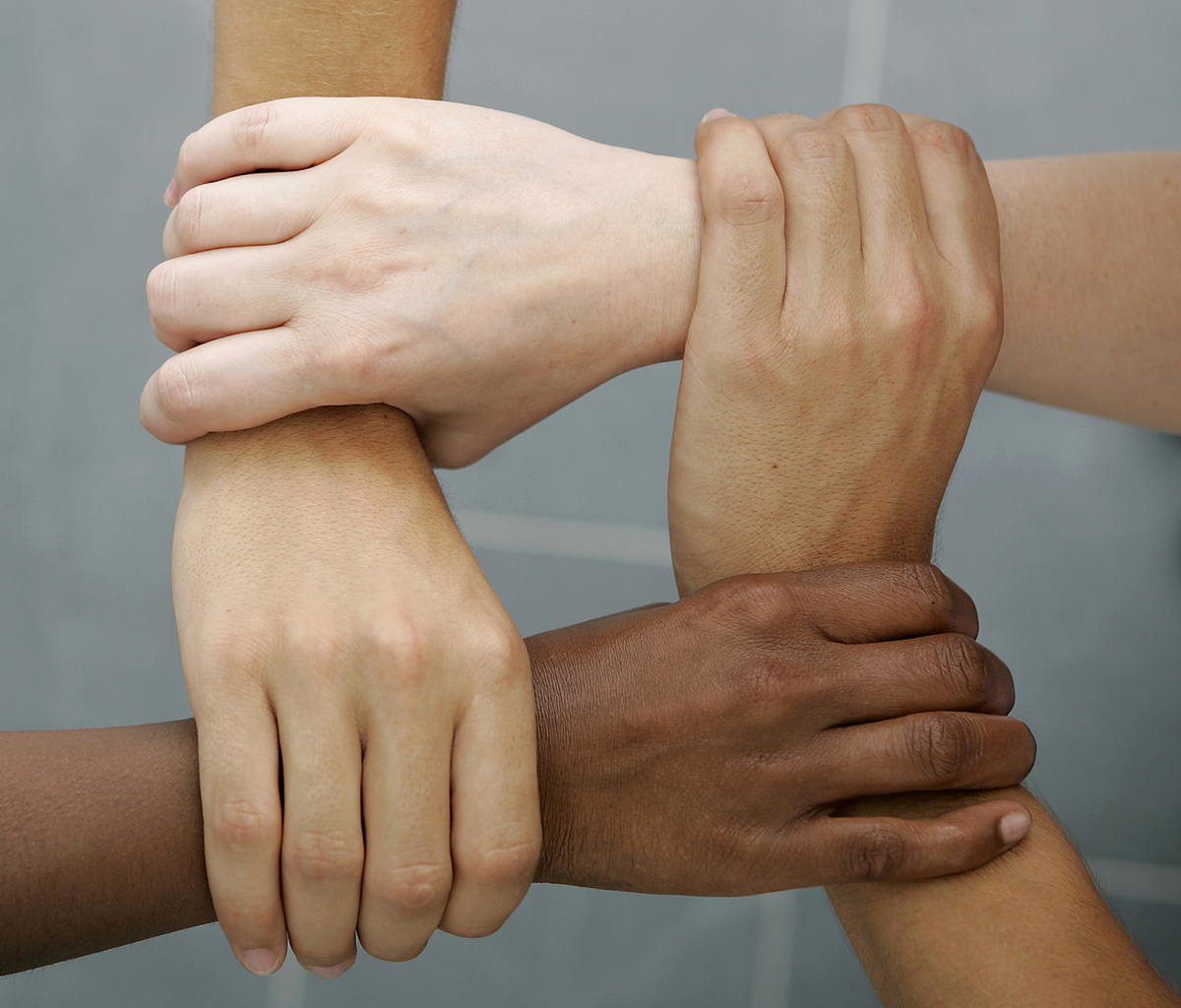 Four hands of people of varying skin tones joining together by holding onto the wrist of the hand in front of it.