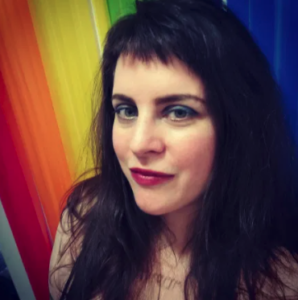 Picture of a white woman with straight black hair and blue eye makeup and red lipstick standing in front of a rainbow flag