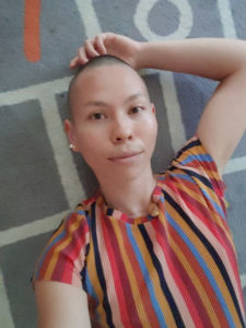 Picture of a nonbinary person of Asian descent in their 20-30s, lying on a grey floor with white square stripes looking directly up at the camera, with short buzzed hair and wearing a colorful, striped shirt