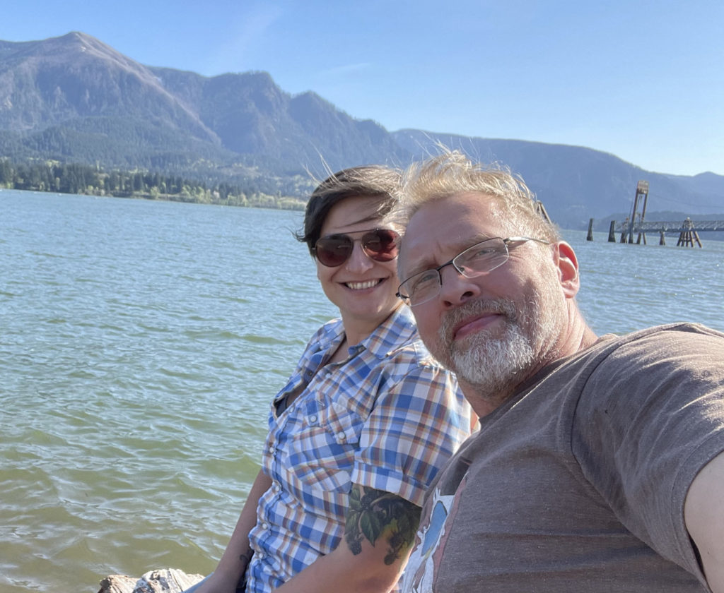 Selfie of a smiling Allison Moon, a white woman with short auburn hair wearing a blue, white and orange checkered shirt and sunglasses, and Reid Mihalko, a blonde, grey bearded, eyeglass-wearing white guy in his 50s, sitting by a rivers edge on a sunny day with the Oregon Gorge in the background