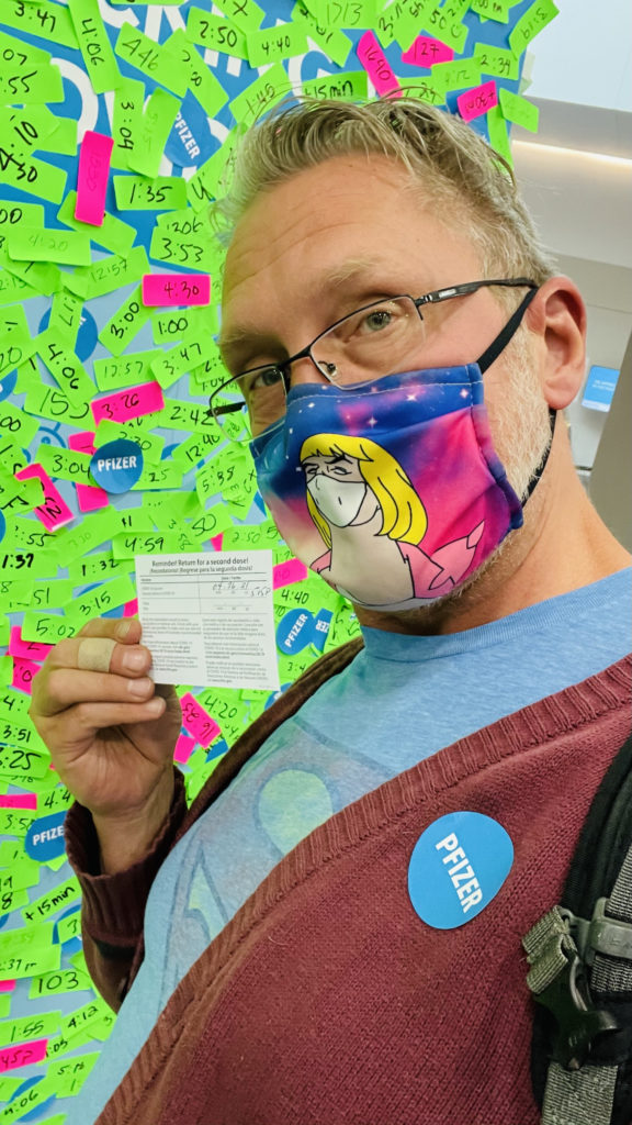 Image of a blonde, white man wearing eyeglasses and a colorful a He-Man meme printed cloth COVID mask holding up his vaccination card and wearing a blue Pfizer sticker on his burgundy sweater