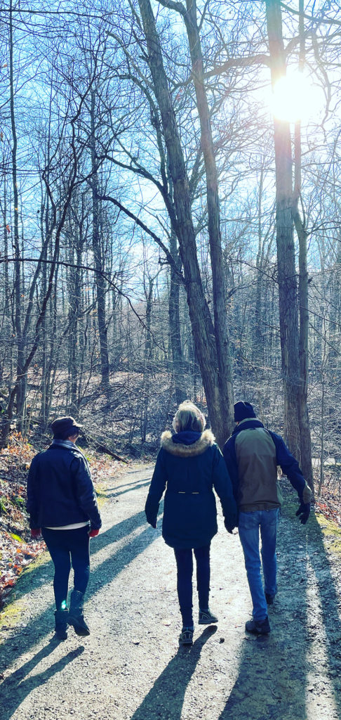 photo of three people walking in the forest on a cold winters day as the setting sun shines through the bare trees and streams across their thick jackets and winter hats. Two of the people are holding hands. 