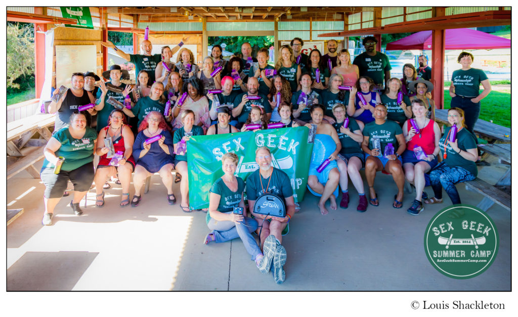 Group photo of 45+ diverse collection of adults from many races, ages, and genders, most of them wearing green, Sex Geek Summer Camp t-shirts and holding purple water bottles for Sybian vibrators. In front of the group, a white man and woman sit on the ground holding a black Sybian vibrator in their laps.