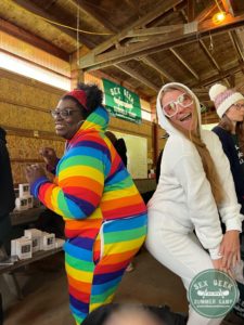 Sex Geek Summer Camp campers wearing onesies and hamming it up for the camera - one camper, a happy black woman in her 30-40s, is wearing a rainbow onesie and the other is a white woman wearing pink sunglasses and a white fuzzy onesie with a hoodie.