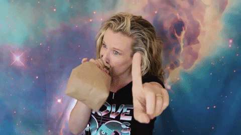 GIF of a blonde woman breathing into a paper bag and hold out her hand in a "Wait a moment" gesture