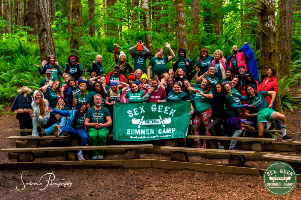 Sex Geek Summer Camp 2022 group photo of approx 55 happy, silly, and colorful humans posing for a group photo in the forest of tall pinetrees