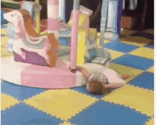 little girl girl - seemingly passed out - being dragged on the floor while holding onto a unicorn merry-go-round