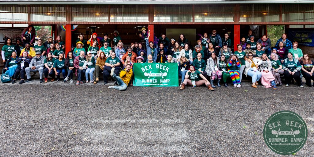 Group photo from 2023's Sex Geek Summer Camp with approximately 85 happy, colorful and diverse adults posing for a photo in front of an outdoorsy barn-type structure