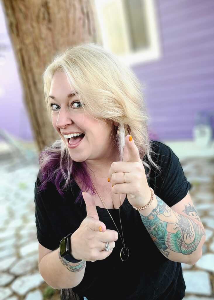 Headshot for Ellecia Paine. A woman with bleach blonde hair with purple tips. She is looking a the camera with her head slightly to the right, an open mouth smile, and finger guns pointed at the camera