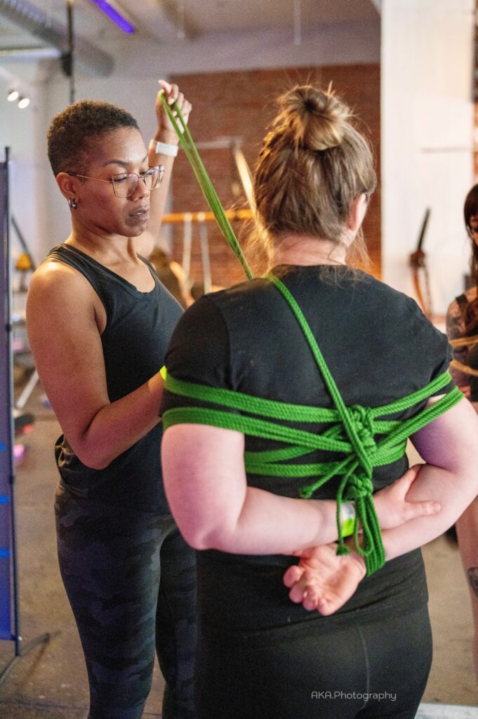 Black woman in a black tank top doing Shibari rope harness tie using green rope on a white woman wearing a black shirt with here arms and hands tied behind her back. 