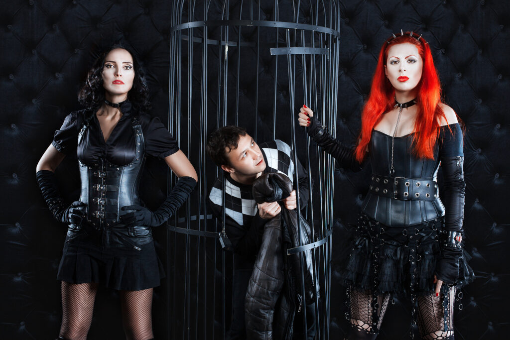 Two women dominatrix dressed in leather, latex, and corsets letting a shy man out of a steel cage